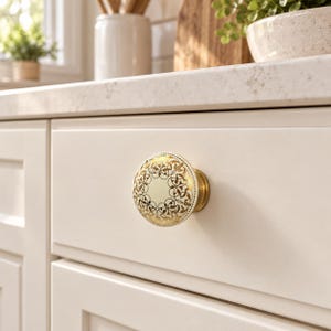 Può includere: Un pomello rotondo per mobile con un centro in ceramica color crema e un motivo a filigrana in oro metallizzato sul bordo. Il pomello ha una base in metallo color ottone ed è montato su un frontale di cassetto bianco sporco con finitura opaca.