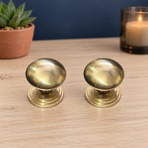 Peut inclure: Une paire de poignées de porte en laiton poli avec un sommet arrondi et lisse et une base circulaire à gradins. La finition métallique reflète la lumière, soulignant la surface incurvée et les anneaux concentriques de la plaque de montage.