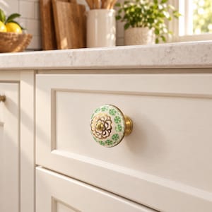May include: A round ceramic cabinet knob featuring a white base with a repeating green floral pattern and a central gold-toned metallic floral design. The knob is mounted on a brass-colored metal base attached to a light-colored wooden drawer front.