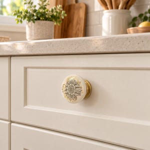 May include: A round ceramic cabinet knob featuring a cream-coloured surface with a detailed black and gold floral mandala pattern. The knob is attached to a brass-toned metal base and is mounted on a light-coloured kitchen drawer front.