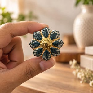 May include: A decorative cabinet knob featuring a floral shape with dark teal petals, white accents, and a central gold-toned metallic circular element. The knob has a scalloped edge and a glossy finish, showcasing a detailed petal pattern.