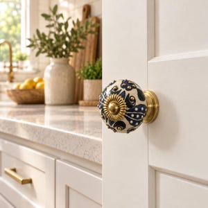 May include: A round ceramic cabinet knob featuring a cream-colored base with a dark blue floral pattern and small gold-toned accents. The center of the knob has a decorative, sunburst-shaped gold metal plate, and it is mounted on a brass base.