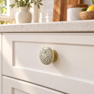 Op de afbeelding: Een ronde meubelknop met een wit keramisch oppervlak voorzien van een zwart geometrisch achthoekig patroon en goudkleurige accenten. De knop is gemonteerd op een messingkleurige metalen voet als decoratief beslagelement voor meubels.