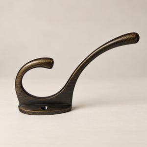 May include: A wall-mounted coat hook with a brushed bronze finish, featuring a curved design with a lower circular loop and an elongated upper arm. The base includes a single pre-drilled screw hole for secure installation on flat surfaces.