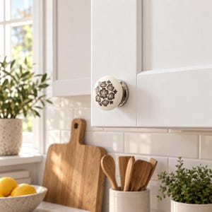 May include: A round white ceramic cabinet knob featuring a dark gray floral pattern with a metallic silver base, mounted on a white cabinet door. The knob has a smooth, glossy finish and a circular shape with a centered decorative flower design.