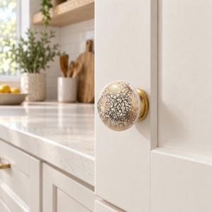 May include: A round cabinet knob featuring a white ceramic base with a detailed gold metallic floral pattern. The knob is mounted on a white cabinet door and includes a circular gold-toned metal base plate that connects it to the cabinet surface.
