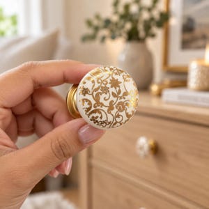 Könnte beinhalten: Ein runder Möbelknopf mit einer weißen Keramikoberfläche, die mit einem metallisch goldenen Blumenrankenmuster verziert ist. Der Knopf hat einen goldfarbenen Metallsockel und eine glatte, kreisförmige Vorderseite mit detaillierten botanischen Motiven.
