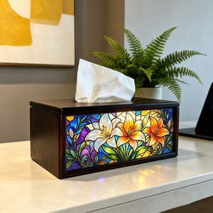 May include: A rectangular dark wood tissue box cover featuring a stained glass panel on the front. The glass displays a vibrant floral pattern with white, orange, and yellow lilies set against blue and purple foliage. A white tissue is visible.