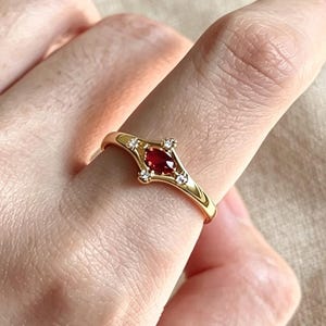 May include: A gold ring featuring a central oval-shaped red gemstone set horizontally. The band includes four small clear stones arranged in a diamond-like pattern around the center, with two stones positioned above and below the red gem.