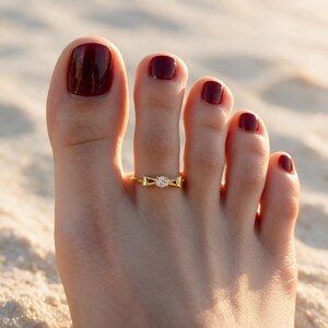 May include: A gold-toned toe ring featuring a central bow-shaped design with a small, clear, round-cut gemstone set in the middle. The ring is worn on the second toe, which has dark red nail polish applied to the toenail.