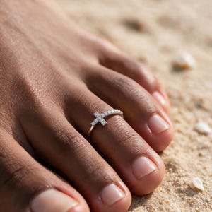 Könnte beinhalten: Ein silberfarbener Zehenring mit einem kleinen Kreuz-Design, das mit klaren, funkelnden Steinen entlang des Rings und auf dem Kreuz besetzt ist. Der Ring wird am zweiten Zeh getragen und zeigt eine feine metallische Oberfläche und Form.