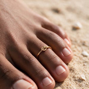 Puede incluir: Un anillo para el dedo del pie en tono dorado con un diseño de nudo central. El anillo es una banda circular delgada que rodea el dedo, mostrando un acabado metálico pulido y un detalle de nudo simple y simétrico en el centro de la banda.