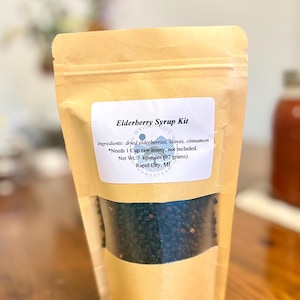 Könnte beinhalten: Ein brauner Standbeutel mit einem Holunderbeeren-Sirup-Set. Das Etikett listet getrocknete Holunderbeeren, Nelken und Zimt als Zutaten auf, mit einem Nettogewicht von 97 Gramm. Ein Sichtfenster zeigt die dunklen, getrockneten Beeren im Inneren.