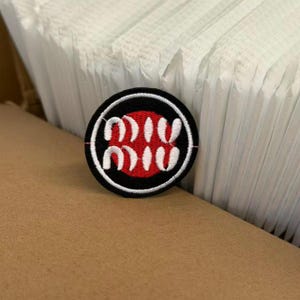May include: A circular embroidered patch featuring a black border and a red center. The white text "miu miu" is stitched in a stylized font across the middle of the red circle. The patch has a white stitched outline around the black border.