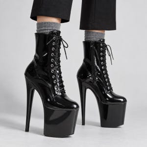 May include: Black patent leather ankle boots featuring a high platform sole, a very tall stiletto heel, and a front lace-up closure with silver-toned eyelets. The boots include a side zipper and are worn with grey ribbed socks and black trousers.