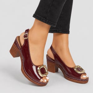 May include: A pair of dark burgundy patent leather slingback sandals featuring a chunky block heel with a wood-grain texture, an open-toe design, and a large, polished gold-tone square buckle accent on the front of each shoe with a thin ankle strap.