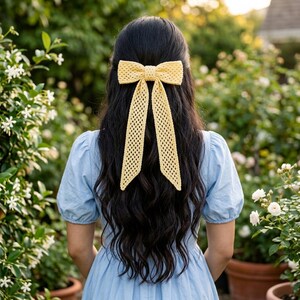 May include: A pale yellow crochet hair bow with a textured, open-knit pattern and two long, dangling ribbon-like tails. The bow is secured at the center with a matching knotted band, creating a structured yet flexible accessory for hair.