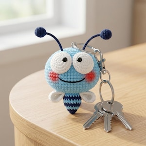 May include: A light blue crocheted bee keyring featuring a round head with large white eyes, black pupils, and pink cheeks. The body has dark blue stripes, small white wings, and two dark blue antennae. A silver metal keyring holds three keys.