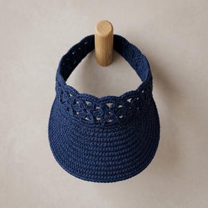 May include: A dark navy blue crochet visor featuring a structured, solid brim and a decorative band composed of repeating floral-inspired openwork motifs. The accessory is crafted from a dense, textured yarn and maintains a circular shape.
