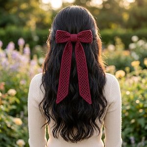 May include: A dark red crochet hair bow with a textured, open-knit pattern. The bow features a central knot and two long, dangling ribbon-like tails that extend downward, secured in long, dark, wavy hair above a cream-colored ribbed long-sleeve top.