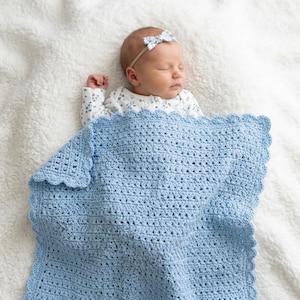 Può includere: Una coperta per neonati all'uncinetto color azzurro con bordo smerlato e motivo a punti in rilievo. La coperta è drappeggiata sopra un neonato che dorme, che indossa un body bianco a maniche lunghe con stampa floreale blu e fascia abbinata.