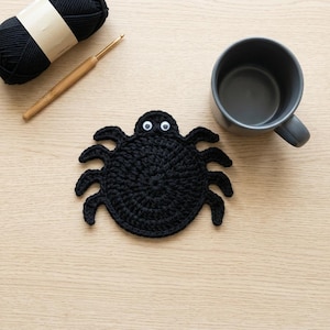 Pode incluir: Um porta-copos de crochê preto em forma de aranha com corpo redondo, oito pernas e dois olhos móveis brancos. O porta-copos está posicionado perto de um novelo de lã preta, uma agulha de crochê de madeira e uma caneca de cerâmica cinza-escura.