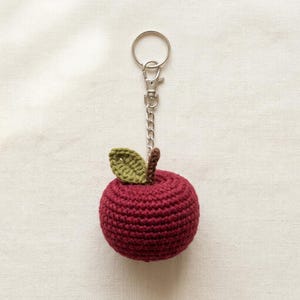 May include: A round, dark red crocheted apple keychain featuring a small, textured green leaf and a short brown stem. The apple is attached to a silver-toned metal split ring by a short chain, providing a secure way to hold keys or accessories.