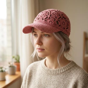 May include: A dusty rose baseball cap crafted from crocheted yarn featuring an intricate floral motif pattern across the crown and a solid, tightly stitched brim. The cap is constructed with a textured, open-work design throughout the main body.