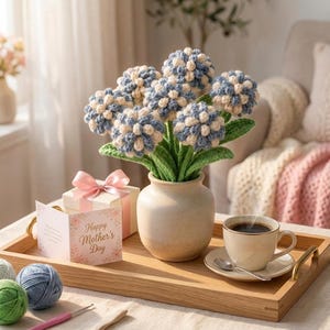 May include: A bouquet of crocheted blue and cream flowers in a beige ceramic vase, accompanied by a gift box with a pink ribbon, a Happy Mother's Day card, a cup of coffee on a saucer with a spoon, and several balls of yarn on a wooden tray.