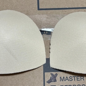 Peut inclure: Deux coussinets de soutien-gorge en tissu beige, également appelés inserts de soutien-gorge, sont présentés sur une surface brune. Les coussinets sont ronds et ont une surface texturée.