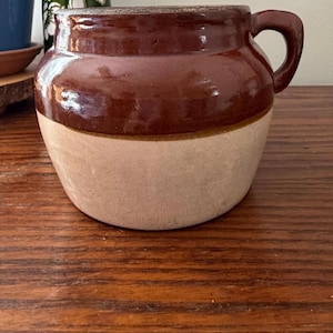 May include: A stoneware crock featuring a two-tone design with a glossy dark brown glazed upper section and a matte beige lower section. The vessel has a wide, rounded body, a thick rim, and a single sturdy handle attached to the side.