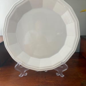May include: A round, off-white ceramic plate featuring a faceted, multi-sided rim design. The plate has a smooth, glossy finish and is displayed upright on a clear, decorative plastic stand, showcasing its geometric border and clean surface.