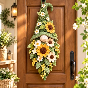 May include: A crochet gnome wall hanging featuring a tall green pointed hat decorated with small white flowers. The gnome has a round beige nose and a beard composed of large yellow sunflowers, white daisies, green leaves, and small yellow berries.