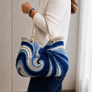 Puede incluir: Un bolso tote grande con construcción de ganchillo y patrones de remolinos en tonos azul marino, azul claro y crema. El bolso tiene dos asas de color crema sujetas con anillos de metal dorado y una abertura superior ancha y estructurada.