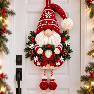 May include: A crocheted gnome wall hanging featuring a red pointed hat with white snowflake patterns and a white pom-pom. The gnome has a white beard, holds a green wreath with red berries and a bow, and has dangling legs with red round feet.