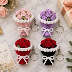 May include: Four crochet flower bouquet keychains in pink, purple, red, and black. Each keychain features a cluster of crocheted roses with green accents, a white ruffled border, and a white crocheted bow, attached to a silver metal keyring.