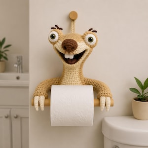 May include: A crocheted sloth-like character wall-mounted toilet paper holder. The figure is light brown with a dark brown nose, two white teeth, and large eyes. It holds a wooden rod supporting a roll of white toilet paper against the wall.