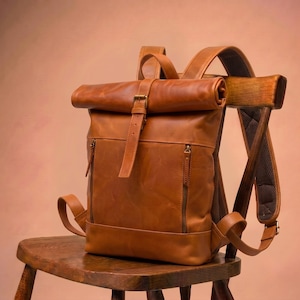 Puede incluir: Una mochila de cuero color canela con cierre enrollable, una correa vertical con hebilla metálica, dos bolsillos frontales verticales con cremallera y tiradores de cuero a juego, y correas ajustables con acolchado de malla marrón.
