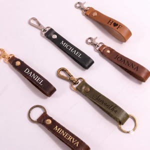 May include: Six leather keychains in various colours including black, brown, olive green, and tan. Each keychain features a metal clasp or ring, a metal rivet, and a personalised name or initials engraved on the leather strap: H heart F, MICHAEL, JOANNA, DANIEL, Samantha, and MINERVA.