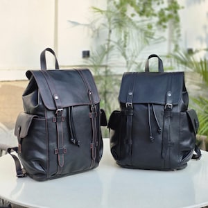Puede incluir: Dos mochilas negras con asas superiores y correas ajustables. Cada bolsa cuenta con una solapa con cierres de hebilla, un compartimento principal con cordón y dos bolsillos laterales. La mochila de la izquierda tiene costuras rojas.
