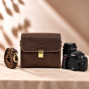 Peut inclure: Un sac photo en cuir marron foncé de forme rectangulaire, doté de surpiqûres blanches contrastantes et d'un fermoir à pression en métal doré. Le sac est accompagné d'une bandoulière amovible assortie avec des ferrures dorées et un appareil Nikon D3200.