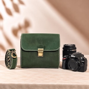 Pode incluir: Uma bolsa para câmara em couro verde escuro com formato retangular, costuras brancas em contraste e um fecho de pressão em metal cor de latão. A bolsa é acompanhada por uma alça de ombro em couro verde a condizer e uma câmara Nikon D3200.