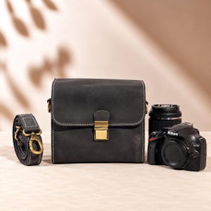 May include: A charcoal grey leather camera bag with white contrast stitching and a brass-toned metal clasp closure. Beside the bag sits a coiled matching leather shoulder strap with brass hardware and a black Nikon D3200 digital camera body.
