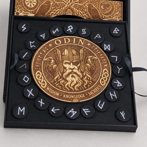 Peut inclure: Une boîte carrée noire contenant une plaque en bois circulaire gravée du portrait d'Odin avec deux corbeaux. La plaque est entourée de jetons circulaires noirs marqués de symboles runiques blancs. Le texte ODIN, WISDOM, KNOWLEDGE, SACRIFICE est visible.