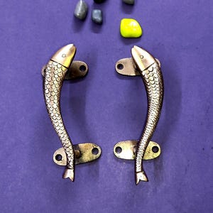 May include: A pair of brass door handles shaped like fish with detailed scale textures. Each handle features two mounting plates with screw holes at the top and bottom. The fish are oriented vertically with their heads pointing upward.
