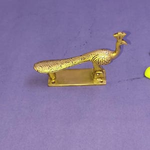 Puede incluir: Un llamador de puerta de latón con forma de pavo real con una cola alargada que presenta detalles de plumas grabados. El ave está montada sobre una placa base rectangular de latón con dos orificios para tornillos visibles.