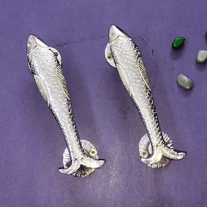 Könnte beinhalten: Zwei silberfarbene Türgriffe aus Metall in Fischform mit detaillierter Schuppenstruktur und geschwungenem Körper. Jeder Griff verfügt über eine kreisförmige Montageplatte am Schwanzende. Daneben liegen vier kleine, glatte, bunte Steine.