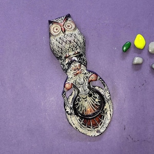 May include: A decorative metal door knocker featuring a detailed owl at the top and a bearded figure below, finished in dark tones with copper highlights. Five small, smooth, multi-coloured stones in yellow, green, and grey are placed nearby.