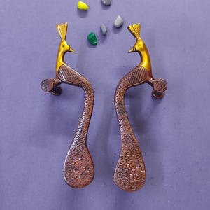 Puede incluir: Un par de manijas de puerta metálicas con forma de pavo real, con cabezas doradas lisas y cuerpos de color cobre texturizados con patrones de plumas detallados. Las manijas están colocadas verticalmente, reflejándose entre sí, junto a pequeñas piedras decorativas.