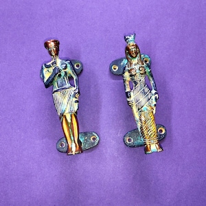 Puede incluir: Dos manijas de puerta metálicas con figuras de estilo egipcio. La manija izquierda muestra una figura con falda, mientras que la derecha muestra una figura con tocado y vestido largo. Ambas tienen placas de montaje con orificios.